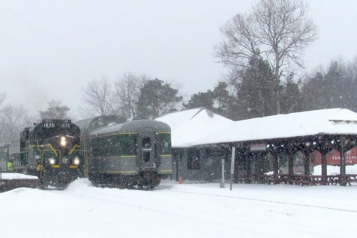 Train arriving at a snowy station with trees in the background, covered in snow.