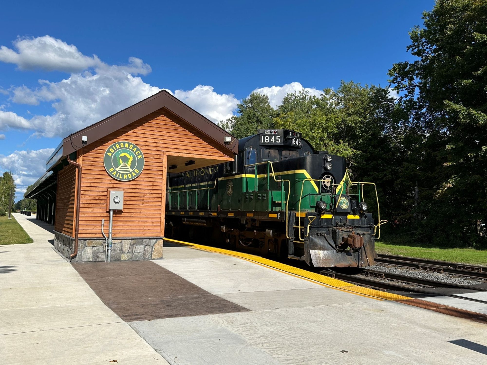 Green train next to wooden building and trees under a blue sky.