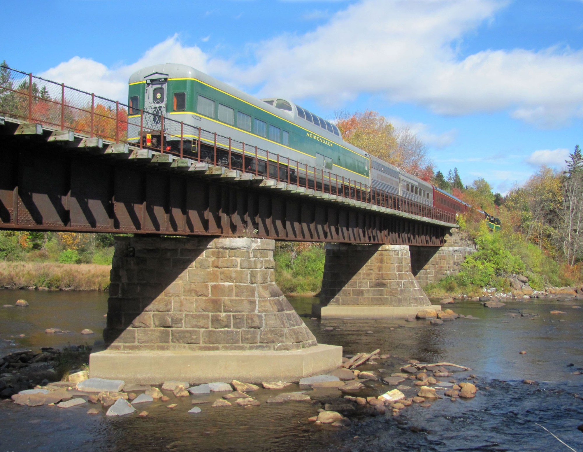 Train crossing a bridge over a river with trees and blue sky in the background.