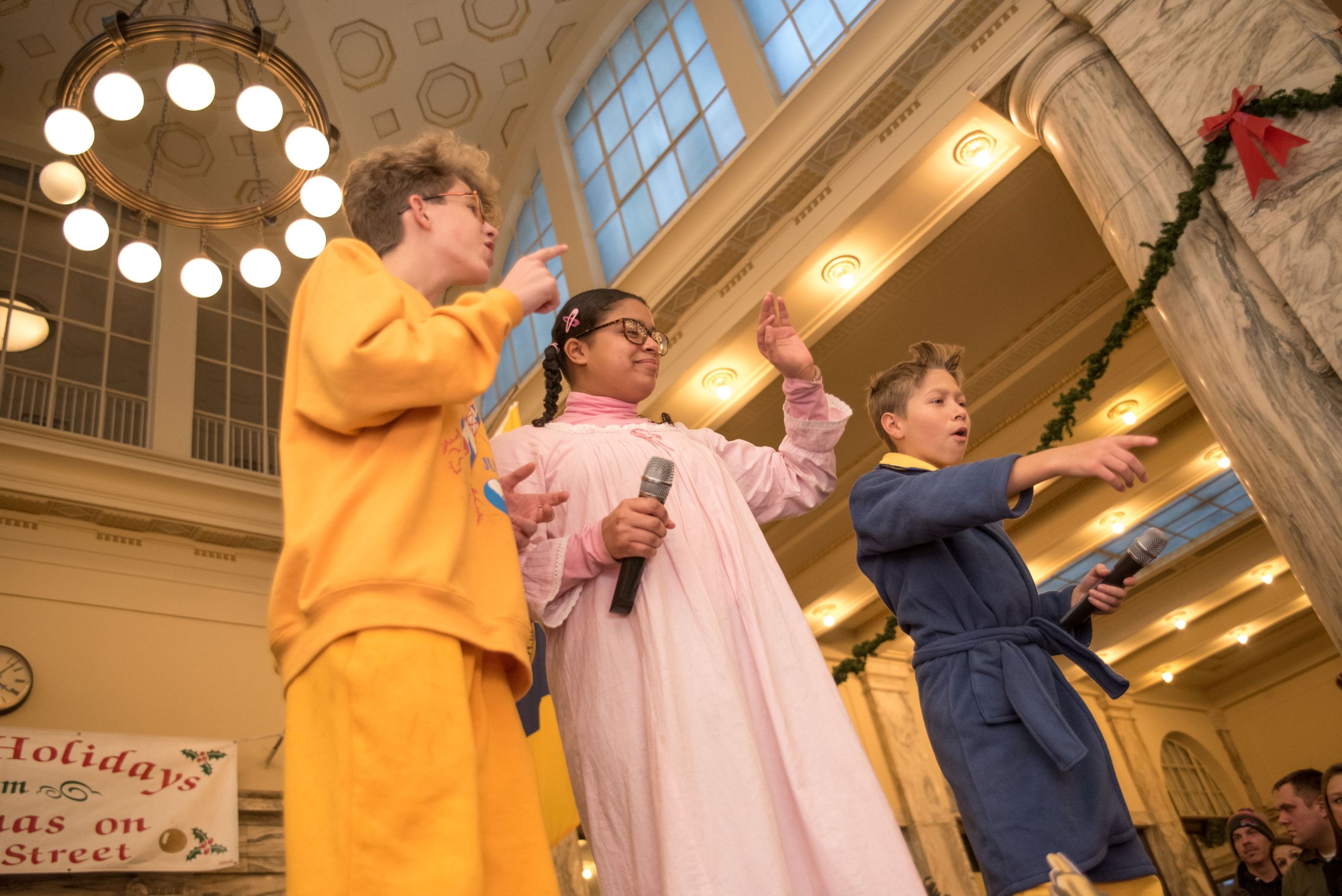 Three children in costumes performing indoors, holding microphones.