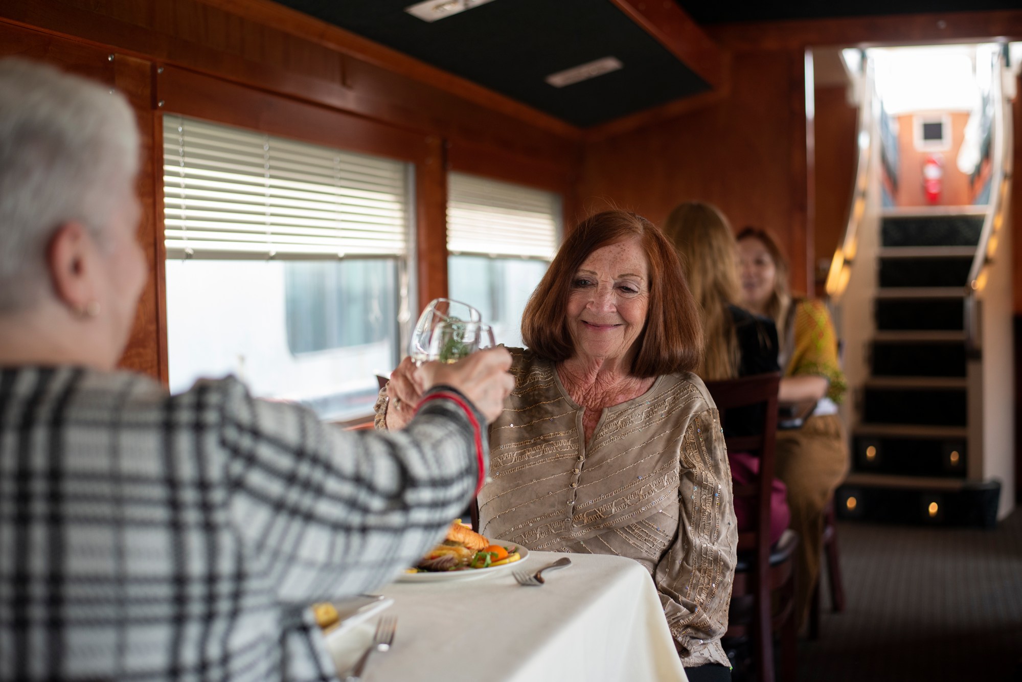 Two women toasting wine glasses at a dining table in a train car.